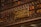 A library wall filled with books on shelves, with a detailed and ornate metal railing in front. Mounted on the rail is a plaque dedicated to Machado de Assis, a Brazilian writer, commemorating his memory.