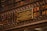 A library wall filled with books on shelves, with a detailed and ornate metal railing in front. Mounted on the rail is a plaque dedicated to Machado de Assis, a Brazilian writer, commemorating his memory.