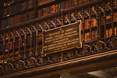A library wall filled with books on shelves, with a detailed and ornate metal railing in front. Mounted on the rail is a plaque dedicated to Machado de Assis, a Brazilian writer, commemorating his memory.