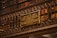 A library wall filled with books on shelves, with a detailed and ornate metal railing in front. Mounted on the rail is a plaque dedicated to Machado de Assis, a Brazilian writer, commemorating his memory.
