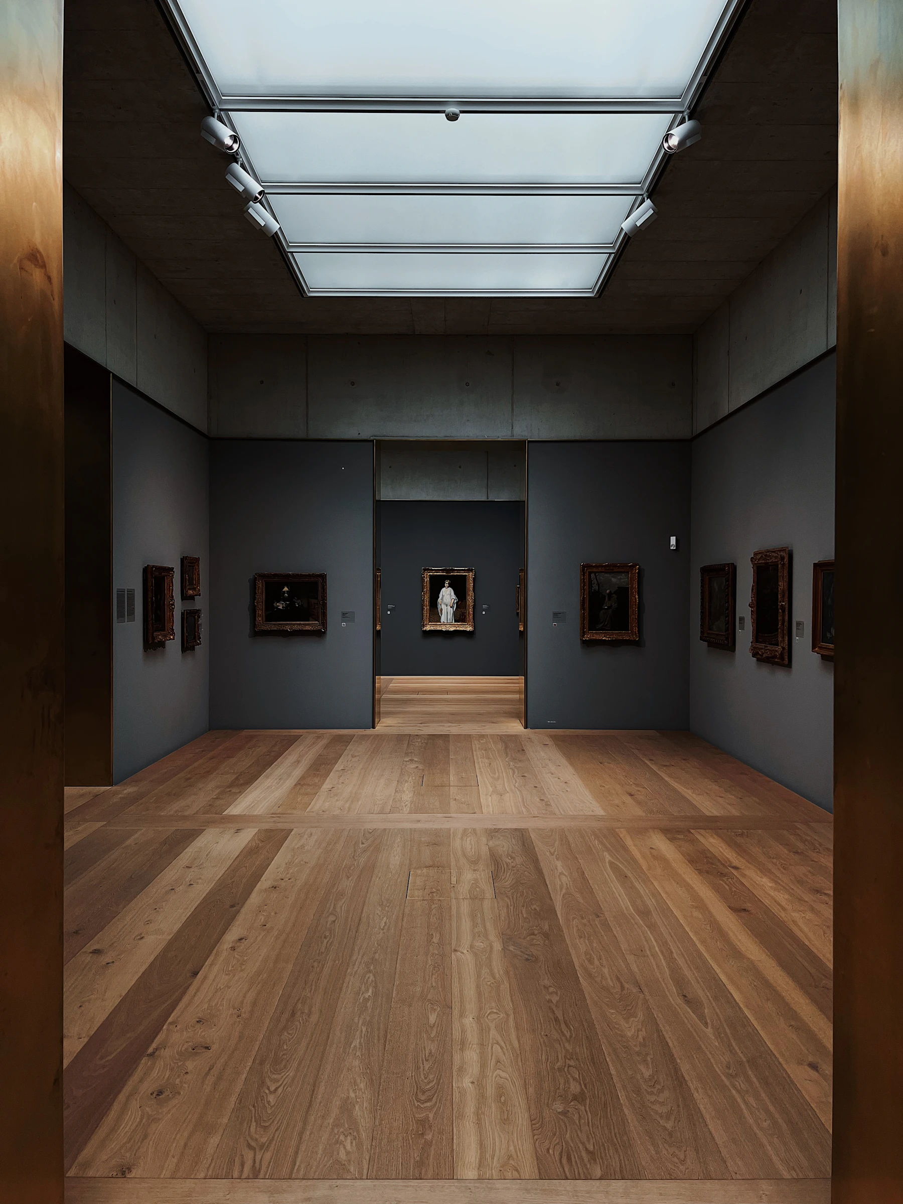An empty gallery has dark walls, framed art, a wooden floor and a large diffused skylight.