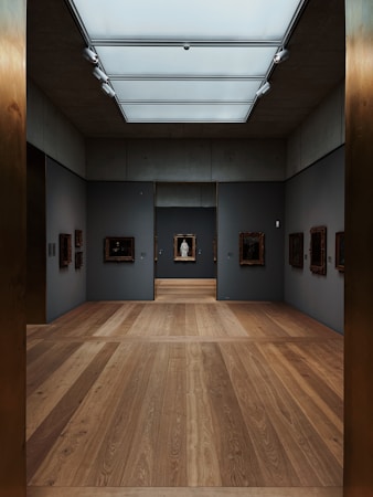 A modern art gallery with dim lighting features paintings displayed on dark gray walls. The room has a sleek design with wooden floors and a skylight that provides soft, natural lighting. The artworks are framed in dark colors, adding contrast to the space.