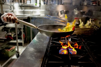 A person wearing gloves is holding a pan with chopped vegetables over an open flame on a stovetop in an industrial kitchen setting. The vegetables appear to be in mid-air as if being tossed. The background shows various kitchen equipment and utensils.
