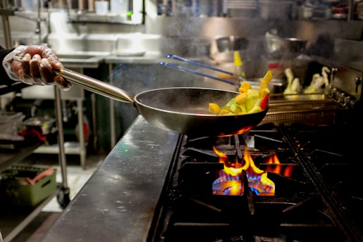 A person wearing gloves is holding a pan with chopped vegetables over an open flame on a stovetop in an industrial kitchen setting. The vegetables appear to be in mid-air as if being tossed. The background shows various kitchen equipment and utensils.