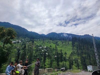 A group of children is gathered outdoors with a backdrop of lush green hills and tall pine trees. The sky is partly cloudy, and the weather appears to be calm. There is also some sort of metal structure, possibly a tower, on the right side. The children seem to be holding sticks or poles and are dressed in casual traditional attire.