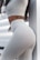 A person wearing a form-fitting, light-colored outfit featuring high-waisted leggings and a crop top. An eagle tattoo is visible on their waist, and the background includes a striped pattern.