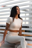 A woman with long dark hair and a tattoo on her arm sits on a chair. She is wearing a fitted, light-colored athletic outfit. The background consists of horizontal white slats and soft furniture.