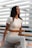 A woman with long dark hair and a tattoo on her arm sits on a chair. She is wearing a fitted, light-colored athletic outfit. The background consists of horizontal white slats and soft furniture.