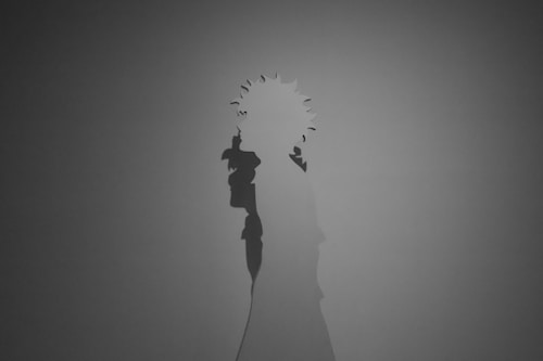 A monochrome image featuring the shadow of a figure with spiky hair, possibly holding an object. The shadow is cast upon a plain surface, creating a simplistic yet intriguing silhouette.