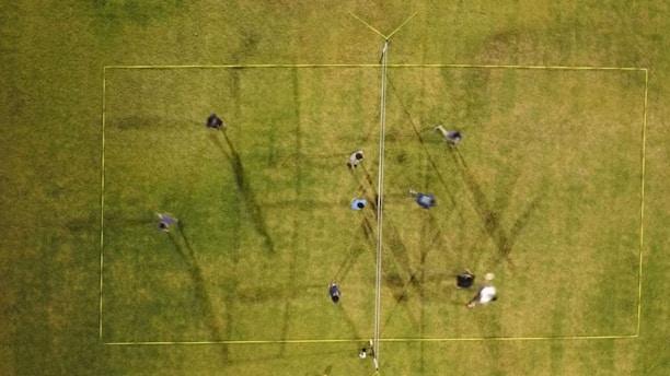 An aerial view of a grass field shows multiple people positioned within the boundaries of a yellow-marked rectangular area. A volleyball net is set up in the center, suggesting a game or practice session. The long shadows cast by the people indicate the sunlight's angle.