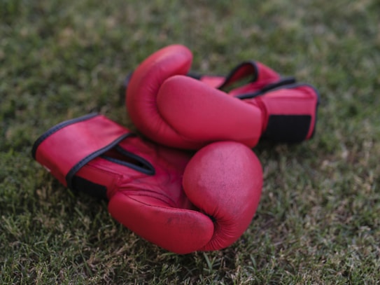 A pair of red boxing gloves lie on a patch of green grass. The gloves are resting with the palms facing upward, and their vibrant red color contrasts sharply against the earthy tones of the grass.