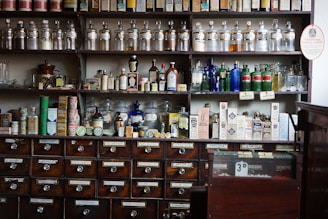 Shelves filled with an array of vintage apothecary bottles and containers, labeled with various substances and remedies. Glass jars and bottles of different shapes and colors, including clear, blue, and amber, are displayed neatly. Below the shelves, a set of wooden drawers, each labeled with different chemical or herbal names, completes the typical old pharmacy look.
