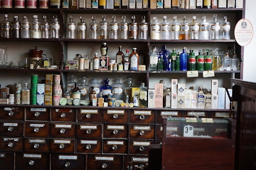 Shelves filled with an array of vintage apothecary bottles and containers, labeled with various substances and remedies. Glass jars and bottles of different shapes and colors, including clear, blue, and amber, are displayed neatly. Below the shelves, a set of wooden drawers, each labeled with different chemical or herbal names, completes the typical old pharmacy look.