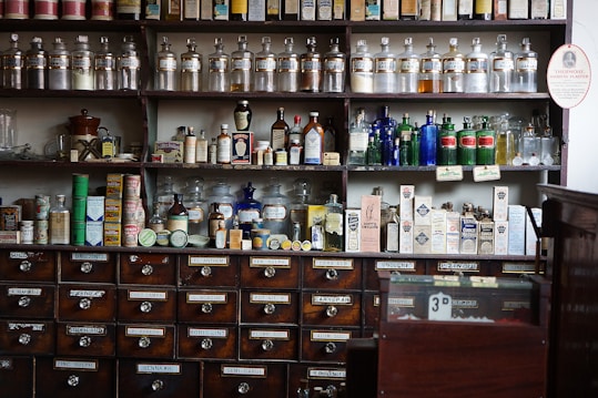 Shelves filled with an array of vintage apothecary bottles and containers, labeled with various substances and remedies. Glass jars and bottles of different shapes and colors, including clear, blue, and amber, are displayed neatly. Below the shelves, a set of wooden drawers, each labeled with different chemical or herbal names, completes the typical old pharmacy look.
