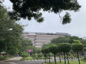 A multi-story building surrounded by lush greenery, with well-maintained trees and shrubs. A pathway lined with black-and-white bollards leads towards the building. In the foreground, a digital signboard is visible among the vegetation.