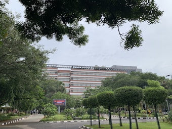 A multi-story building surrounded by lush greenery, with well-maintained trees and shrubs. A pathway lined with black-and-white bollards leads towards the building. In the foreground, a digital signboard is visible among the vegetation.