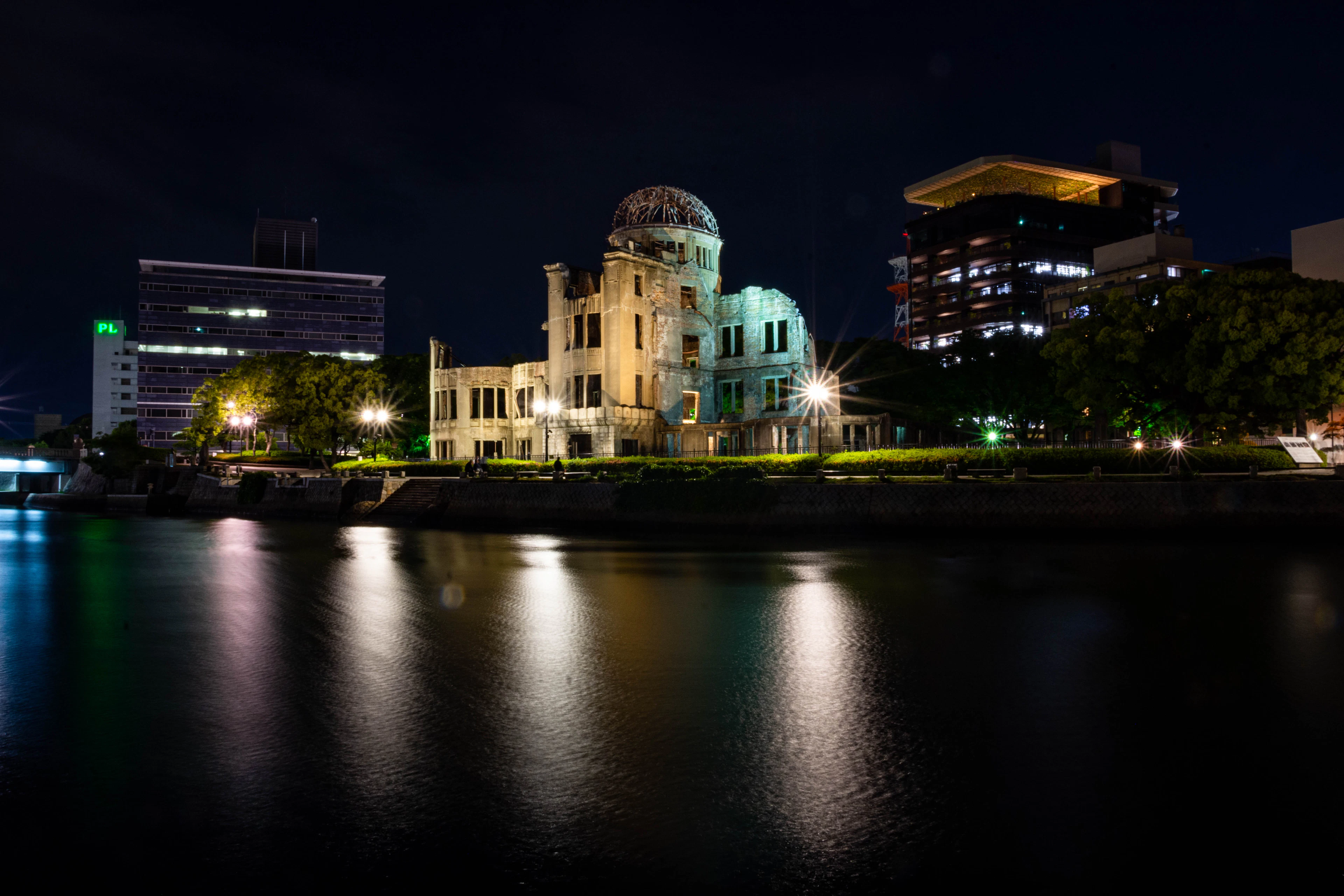 The skeletal dome and gutted brick shell of the Atomic Bomb Dome floodlit at night in Hiroshima's Peace Memorial Park, its light laid in long ribbons across the black water of the Motoyasu river