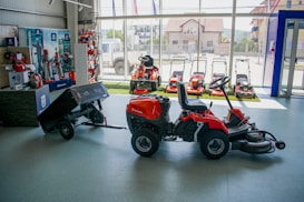A showroom displaying various garden machinery, including several red riding lawn mowers and a variety of power tools. The central focus is a red ride-on mower with an attached cart. The background features a large window with views of buildings outside.