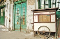 A weathered street food cart labeled 'Gado Gado Nasi' is positioned against an old, deteriorating building with green, wooden doors. The cart has a large wheel at the base and shows signs of wear. The building's façade is marked by peeling paint and exposed bricks.