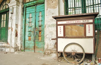 A weathered street food cart labeled 'Gado Gado Nasi' is positioned against an old, deteriorating building with green, wooden doors. The cart has a large wheel at the base and shows signs of wear. The building's façade is marked by peeling paint and exposed bricks.