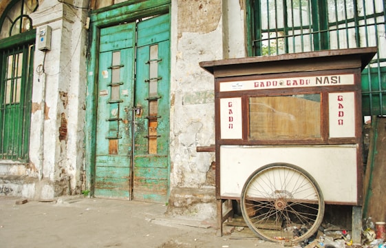 A weathered street food cart labeled 'Gado Gado Nasi' is positioned against an old, deteriorating building with green, wooden doors. The cart has a large wheel at the base and shows signs of wear. The building's façade is marked by peeling paint and exposed bricks.