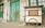 A weathered street food cart labeled 'Gado Gado Nasi' is positioned against an old, deteriorating building with green, wooden doors. The cart has a large wheel at the base and shows signs of wear. The building's façade is marked by peeling paint and exposed bricks.