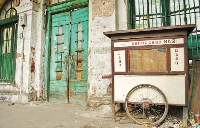 A weathered street food cart labeled 'Gado Gado Nasi' is positioned against an old, deteriorating building with green, wooden doors. The cart has a large wheel at the base and shows signs of wear. The building's façade is marked by peeling paint and exposed bricks.