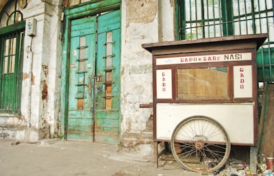 A weathered street food cart labeled 'Gado Gado Nasi' is positioned against an old, deteriorating building with green, wooden doors. The cart has a large wheel at the base and shows signs of wear. The building's façade is marked by peeling paint and exposed bricks.