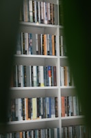 A well-organized bookshelf filled with various books, each with different colored covers and titles in diverse languages. The shelves are part of a white bookcase, and the image is framed by out-of-focus green leaves in the foreground, creating a sense of depth.
