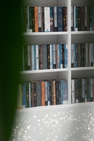 A bookshelf is filled with neatly arranged books, most of which have dark spines with Arabic script. Some sections of the bookshelf are monochromatic, while others vary in color. A blurred green element partially obstructs the view, and soft speckles of light are visible on the bottom portion.