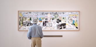 A person wearing a light blue shirt and white hat stands in front of a large, colorful artwork displayed on a gallery wall. The artwork features a complex, abstract scene with various shapes and figures interspersed with vibrant colors.