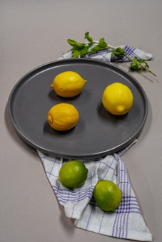 Three ripe lemons are arranged on a dark gray plate, accompanied by two vibrant limes on a blue and white checkered cloth. Fresh mint leaves add a touch of greenery to the composition, contributing to a fresh and natural aesthetic.