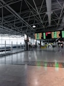 A spacious and modern airport terminal with high ceilings and exposed beams. There are large digital departure boards displaying flight information in bright colors. The terminal features wide open spaces with polished marble floors, a row of benches along the glass windows, and people in the background engaging in various activities.