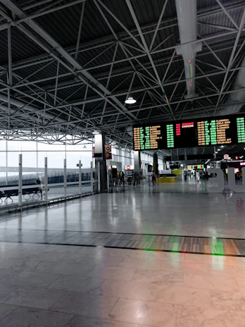 A spacious and modern airport terminal with high ceilings and exposed beams. There are large digital departure boards displaying flight information in bright colors. The terminal features wide open spaces with polished marble floors, a row of benches along the glass windows, and people in the background engaging in various activities.
