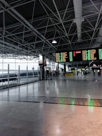 A spacious and modern airport terminal with high ceilings and exposed beams. There are large digital departure boards displaying flight information in bright colors. The terminal features wide open spaces with polished marble floors, a row of benches along the glass windows, and people in the background engaging in various activities.