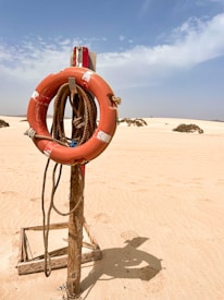 A life preserver hangs on a wooden post secured in the sand of a desert-like landscape. The bright orange of the life ring contrasts with the pale sand and clear blue sky. The area is sparse with a few small bushes scattered in the background.