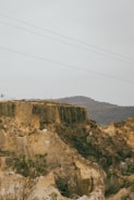A natural landscape featuring steep limestone cliffs with rugged textures. Sparse vegetation grows along the cliff edges and the land appears dry. Hills rise in the background under a cloudy sky, and power lines stretch across the upper part of the scene.