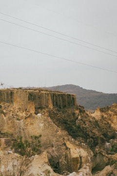 A natural landscape featuring steep limestone cliffs with rugged textures. Sparse vegetation grows along the cliff edges and the land appears dry. Hills rise in the background under a cloudy sky, and power lines stretch across the upper part of the scene.