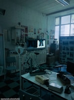 A vintage medical office with retro equipment, including old X-ray images displayed on a lightbox. The room has a checkered black and blue floor, a variety of medical devices, a telephone on a desk, and posters on the walls. Large windows let in natural light.