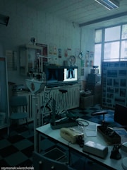 A vintage medical office with retro equipment, including old X-ray images displayed on a lightbox. The room has a checkered black and blue floor, a variety of medical devices, a telephone on a desk, and posters on the walls. Large windows let in natural light.