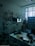 A vintage medical office with retro equipment, including old X-ray images displayed on a lightbox. The room has a checkered black and blue floor, a variety of medical devices, a telephone on a desk, and posters on the walls. Large windows let in natural light.