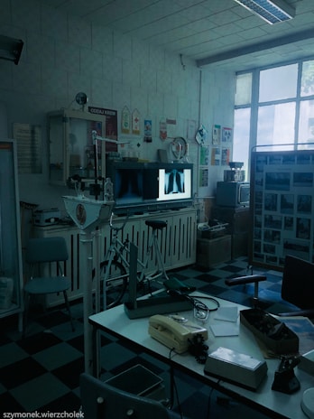 A vintage medical office with retro equipment, including old X-ray images displayed on a lightbox. The room has a checkered black and blue floor, a variety of medical devices, a telephone on a desk, and posters on the walls. Large windows let in natural light.