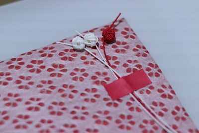 A beautifully wrapped package features a decorative patterned paper with small, red floral designs on a pink background. The package is adorned with three intricate knots in red and white string, finely detailed and placed thoughtfully. A small red strip of tape holds part of the arrangement together.