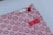 A beautifully wrapped package features a decorative patterned paper with small, red floral designs on a pink background. The package is adorned with three intricate knots in red and white string, finely detailed and placed thoughtfully. A small red strip of tape holds part of the arrangement together.