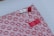A beautifully wrapped package features a decorative patterned paper with small, red floral designs on a pink background. The package is adorned with three intricate knots in red and white string, finely detailed and placed thoughtfully. A small red strip of tape holds part of the arrangement together.