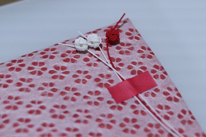A beautifully wrapped package features a decorative patterned paper with small, red floral designs on a pink background. The package is adorned with three intricate knots in red and white string, finely detailed and placed thoughtfully. A small red strip of tape holds part of the arrangement together.