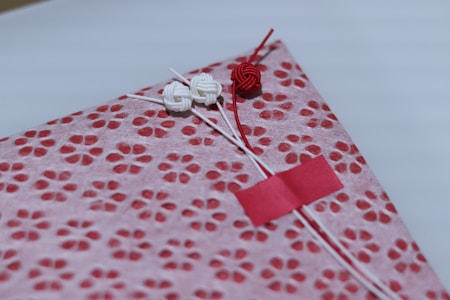 A beautifully wrapped package features a decorative patterned paper with small, red floral designs on a pink background. The package is adorned with three intricate knots in red and white string, finely detailed and placed thoughtfully. A small red strip of tape holds part of the arrangement together.