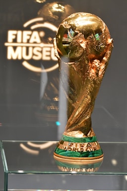A shiny, golden trophy intricately designed with a globe-like top and detailed engravings sits on a glass shelf. The base is highlighted with green and gold, featuring the words 'FIFA WORLD CUP'. The backdrop includes a glass or acrylic panel etched with 'FIFA MUSEUM'.