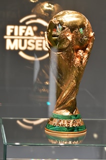 A shiny, golden trophy intricately designed with a globe-like top and detailed engravings sits on a glass shelf. The base is highlighted with green and gold, featuring the words 'FIFA WORLD CUP'. The backdrop includes a glass or acrylic panel etched with 'FIFA MUSEUM'.
