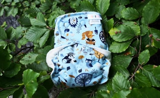 A cloth diaper with a blue background featuring illustrations of woodland animals such as a raccoon and an owl. The diaper is laying on a bed of lush green leaves.